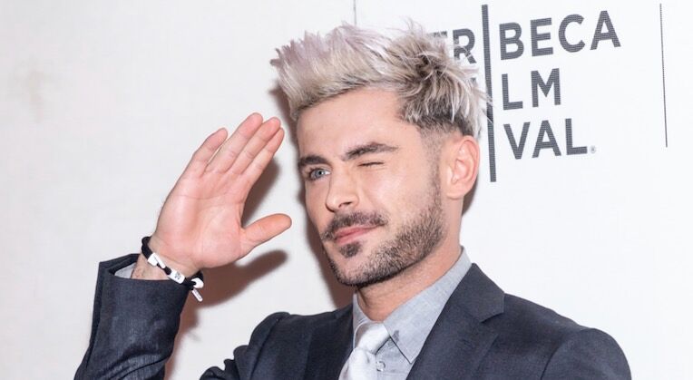 Hollywood actor Zac Efron wears a blazer jacket, undershirt and tie. He has spikey, grey hair and is making a silly face and doing a salute with his hand.