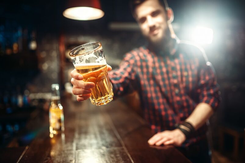 Man in a checkered shirt sitting at a wooden bar holding up a glass of beer.