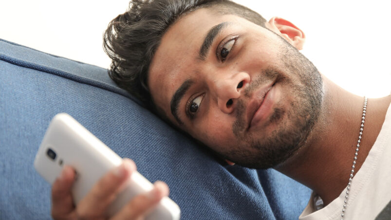 Bearded man lays on his bed and looks at his cellphone.