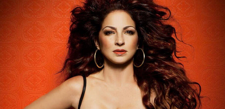 Gloria Estefan with long black hair and large gold hoop earrings standing in front of a red background.