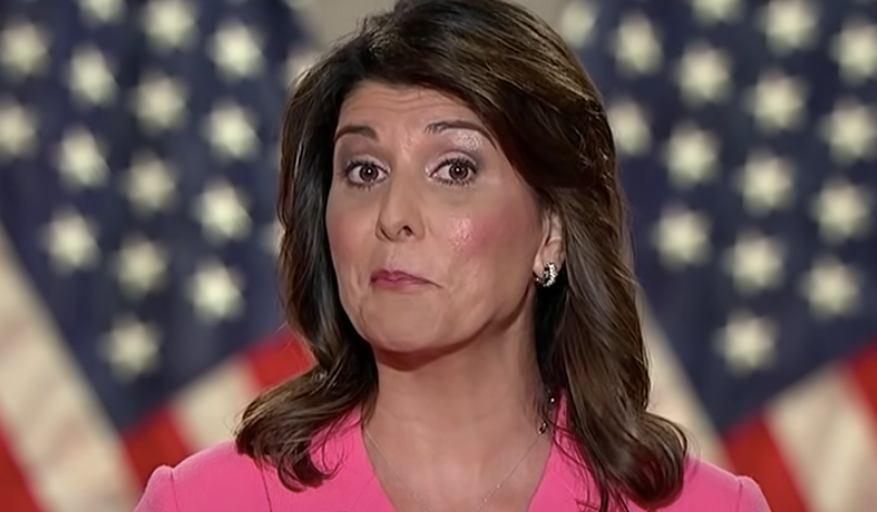Nikki Haley wearing makeup and a pink top in front of an American flag. 