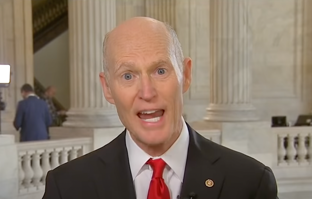 Rick Scott wearing a dark suit jacket with white dress shirt and red tie. 
