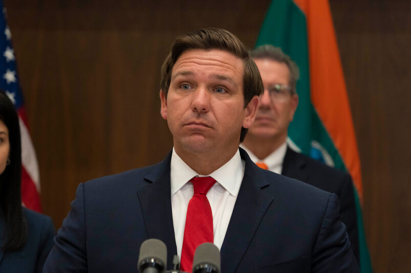 Ron DeSantis standing behind microphones wearing a dark suit jacket, white shirt and red tie.