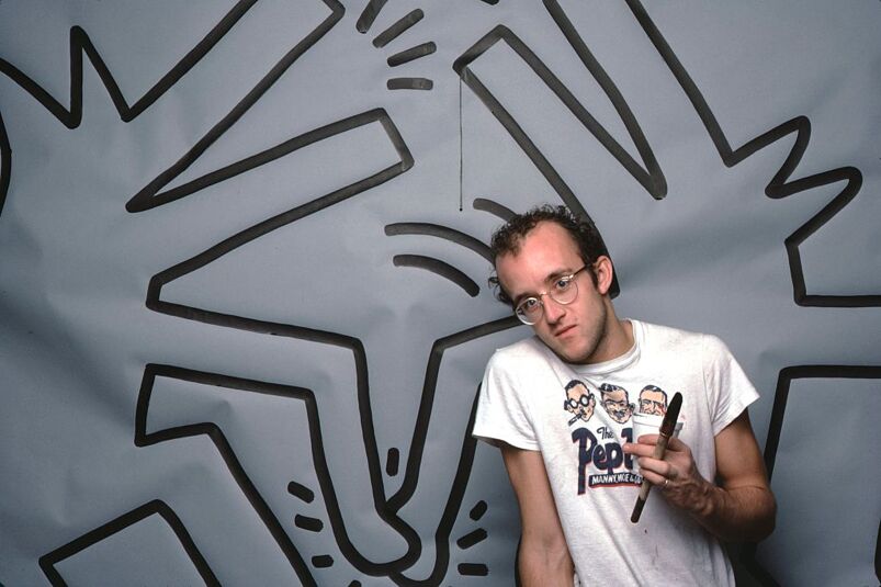 Keith Haring standing in front of his mural holding a paintbrush and cup. 