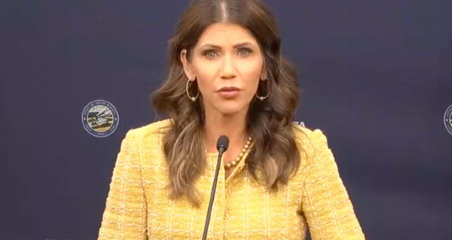South Dakota Governor Kristi Noem standing behind a microphone and wearing a gold business suit 