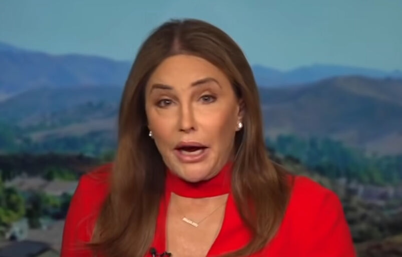 Caitlyn Jenner in a red dress and scarf talking in front of a mountain backdrop