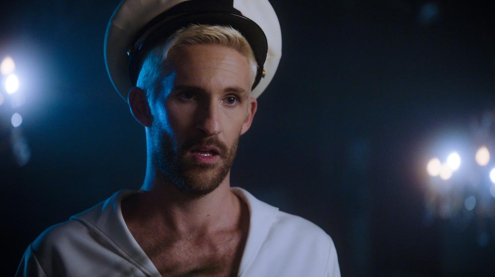 Man with dyed blonde hair is standing in the dark wearing a white sailor's hat.
