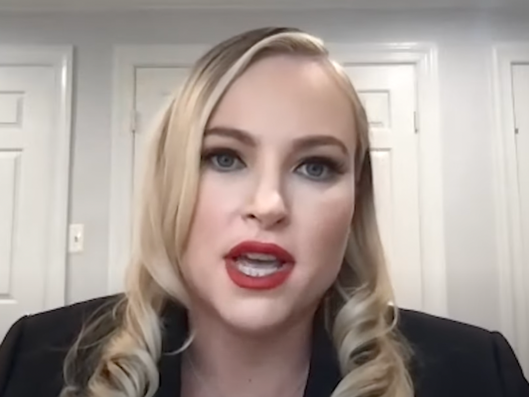 Meghan McCain with red lipstick sitting in front of a white door talking into a camera.