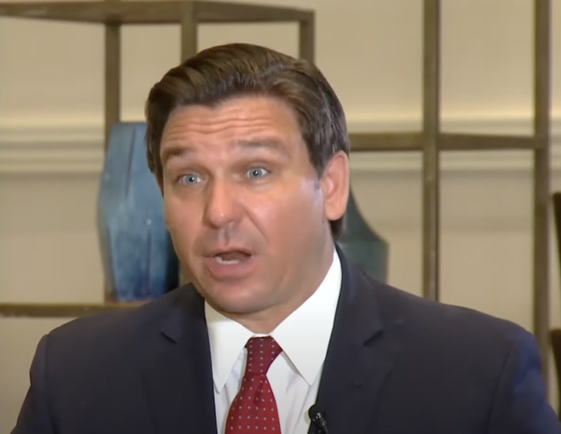 Ron Desantis looking shocked, while wearing a dark suit jacket, white shirt and red tie with white dots.