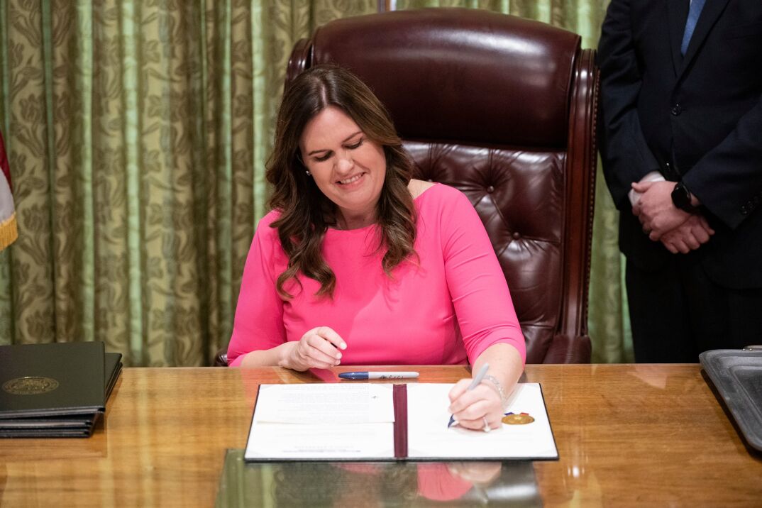 Sarah Huckabee Sanders in a pink dress signing a document at her desk