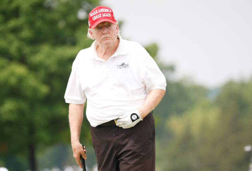 Donald Trump in a white polo shirt and red hat holding a golf  club and looking sad.