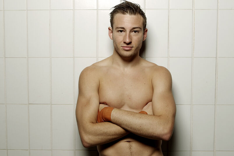 Olympic diver Matthew Mitcham standing against an off-white tile wall shirtless with his arms folded.