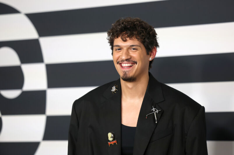 Omar Apollo smiles while wearing a black blazer while on a press line with a black and white backdrop