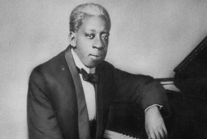 Pianist Tony Jackson sits at a grand piano in a black tuxedo with a bow tie.