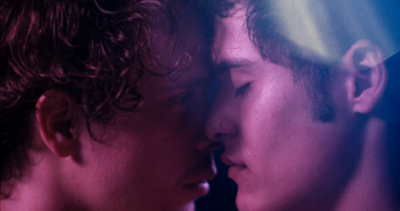 Two men hold their faces close to one another, about to kiss, as they're pathed in a pink-purple light.