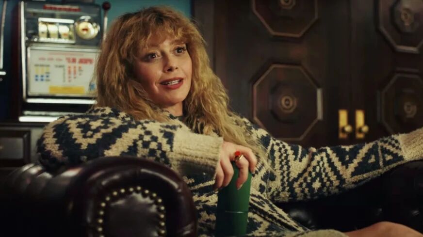 Natasha Lyonne sits in a leather recliner chair while wearing a large grey, patterned cardigan.