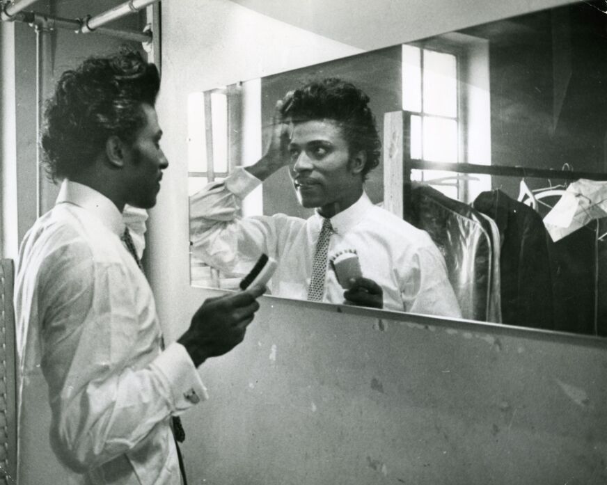Little Richard wears a white shirt and tie as he fixes his pompadour in the mirror