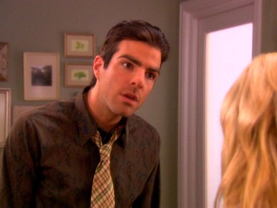 Zachary Quinto, wearing a grey button-up shirt and tie, looks at a blonde woman who is off camera.
