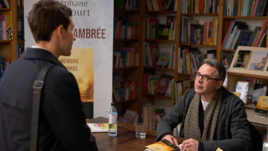 In a book store, and older man in glasses signs a book as he looks at a younger man across the table from him.