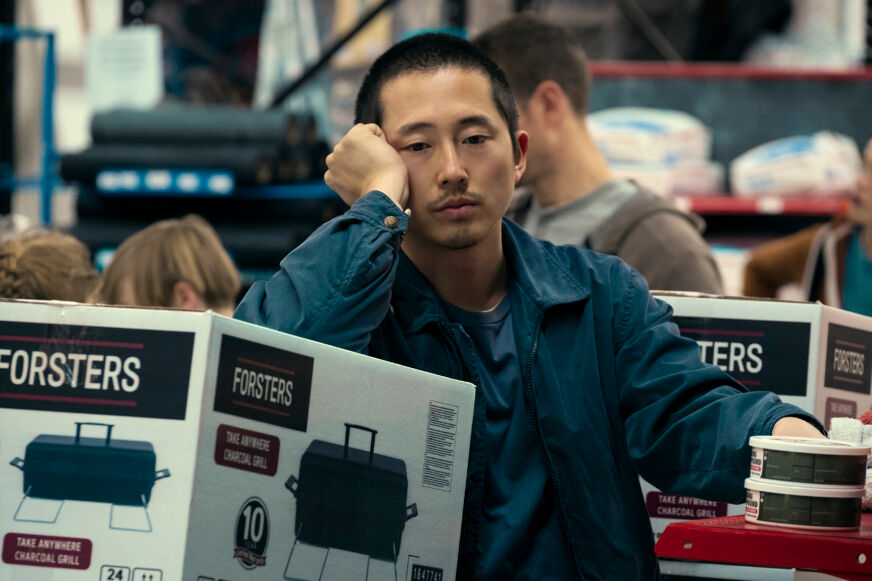 Steven Yeun looks sad in a blue shirt as he leans on a boxed appliance.