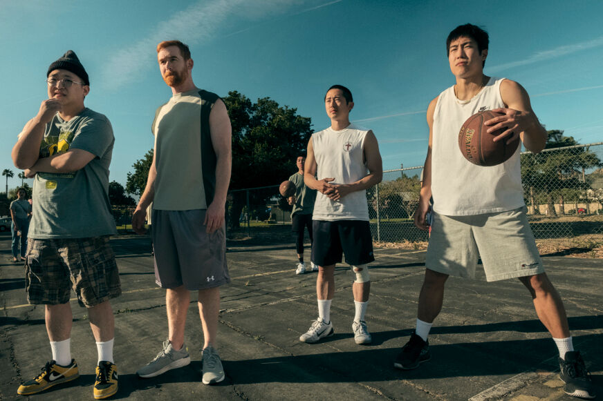 Four men, including Steven Yeun, wear athletic clothes and stand on a basketball court with the blue sky behind them.