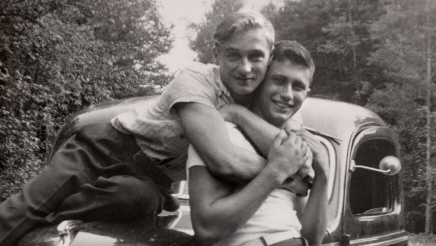 In a black and white antique photo, two young men embrace each other in front of a truck