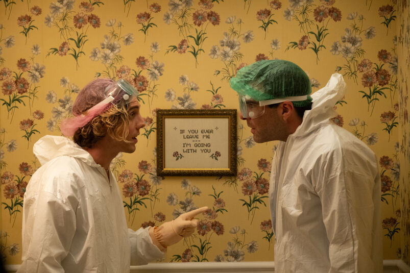 Lukas Gage and Zachary Quinto wear white jumpers, goggles, and hairnets, staring at each other in front of a yellow wall with a flower print.