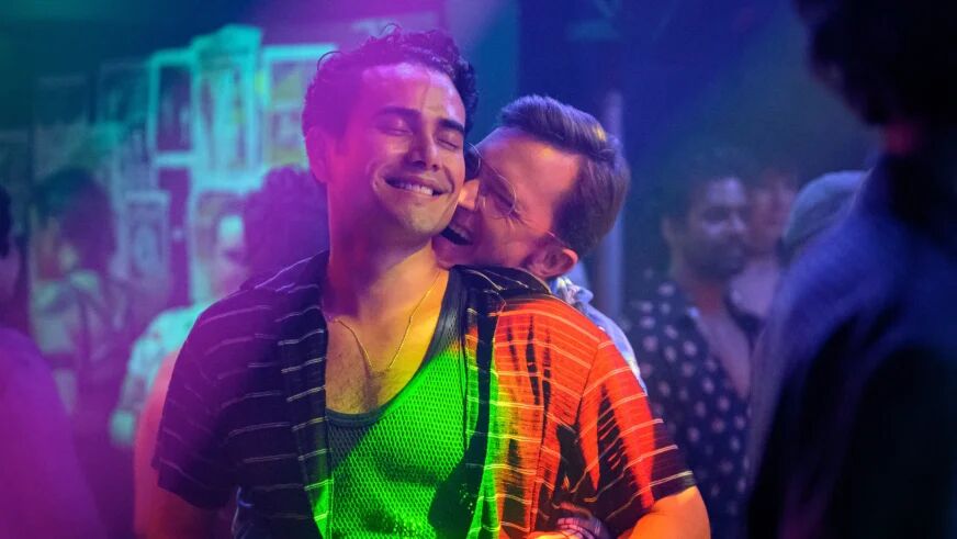 Two gay men dance and smile, colorfully lit in the middle of a dance club