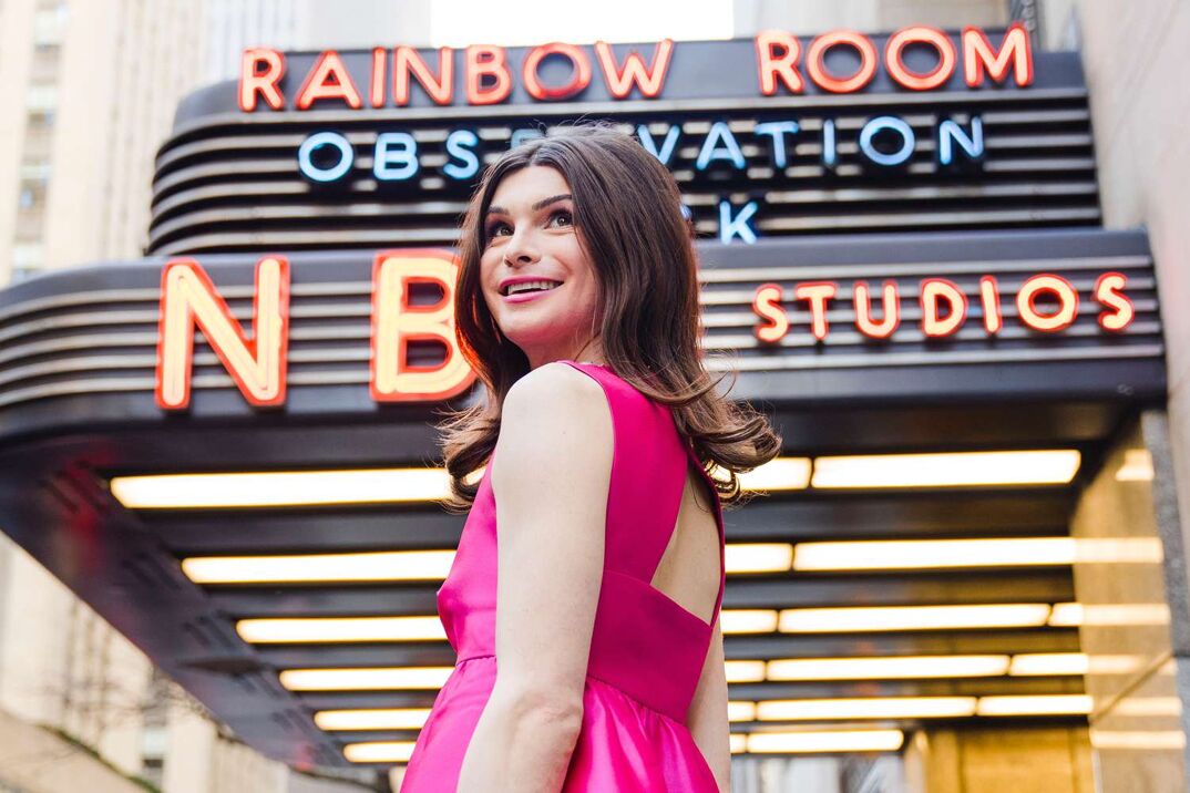 Dylan Mulvaney stands in front of the NBC Studios Observation Deck marquee wearing a pink A-line dress. She looks over her shoulder above the camera, smiling.