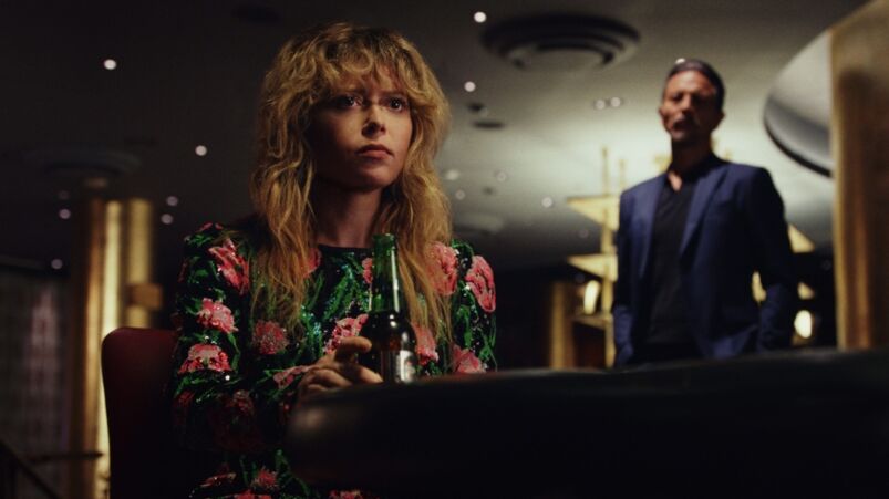 Natasha Lyonne wear a pink flower-covered desk while sitting at a poker table in a dark casino with Benjamin bratt looking over her shoulder