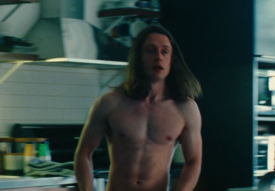 A shirtless and muscled Rory Culkin stands in the middle of a grey kitchen