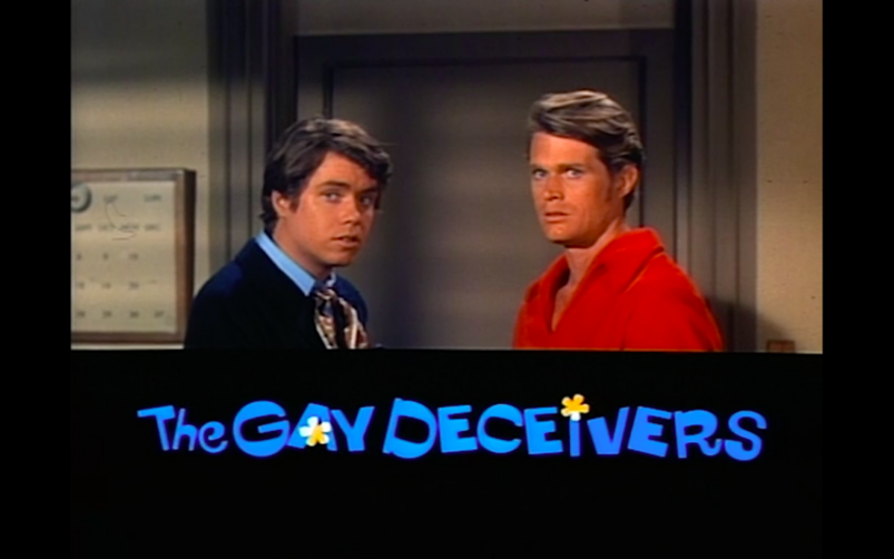 Two men—one in a black coat and one in red—stand in front of a hallway with a title card below them reading, 'The Gay Deceivers'