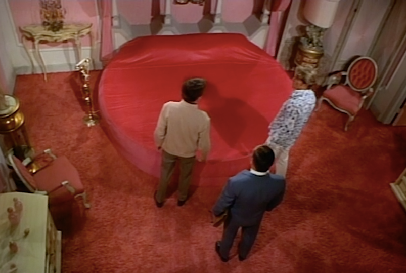 An overhead view of three men in a pink-carpeted room looking at a large, round, red bed