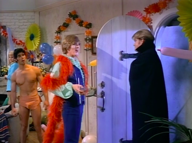 At a decorated costume party, a man in a big red boa greets a man in a black cape at the door as another man in a. small loincloth stands off to the side.