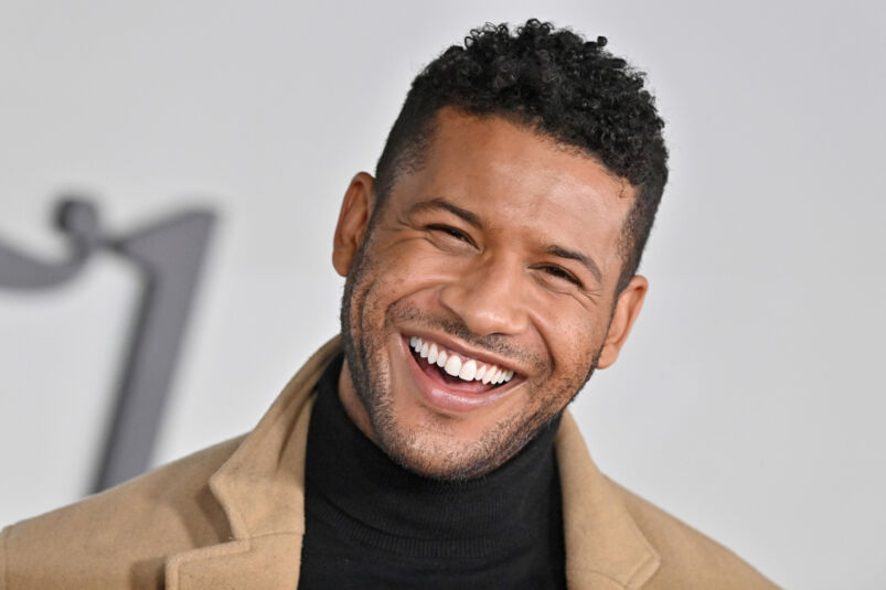 Actor Jeffrey Bowyer-Chapman smiles while wearing a black turtleneck and a tan coat on a red carpet.
