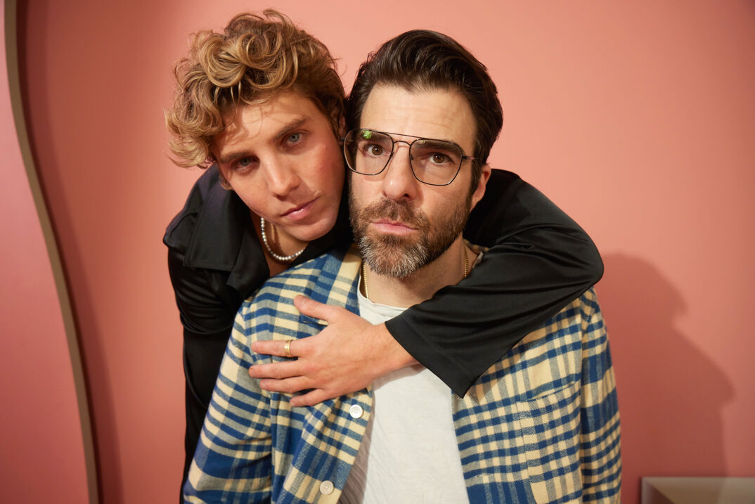 Lukas Gage has his arm Zachary Quinto as the two pose for a photoshoot in a salmon-colored studio.