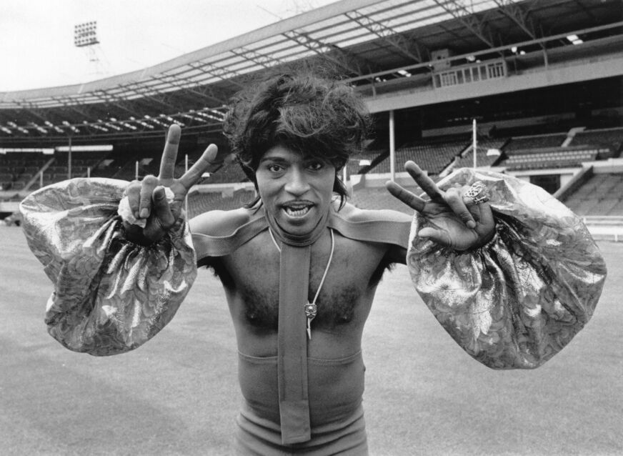 At a large stadium, Little Richard wears a chest-bearing shirt with large sleeves as he flashes two peace signs and sticks his tongue out at the camera.