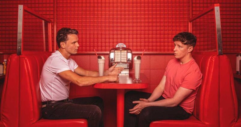 In a bright red diner with red tiling and a red booth, two men sit across from one another looking nervous
