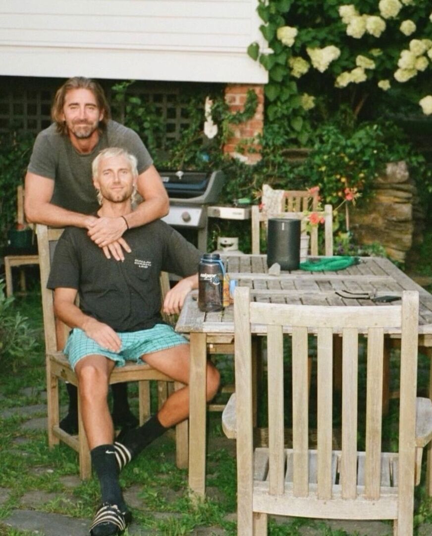 At a picnic table outside, Lee Pace stands behind husband Matt Foley and putss his arms around him.