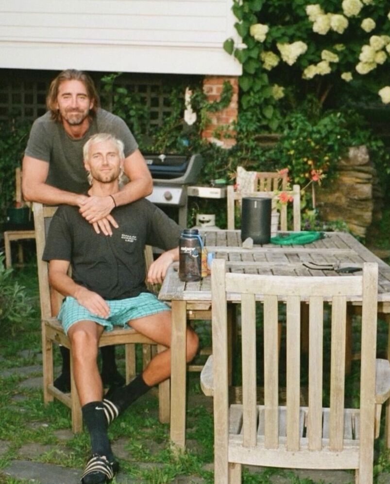 At a picnic table outside, Lee Pace stands behind husband Matt Foley and putss his arms around him.