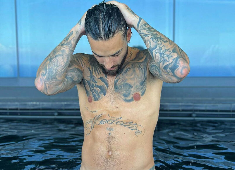 Maluma shirtless in his large swimming pool. He's facing down into the water while his hands slick back his hair.