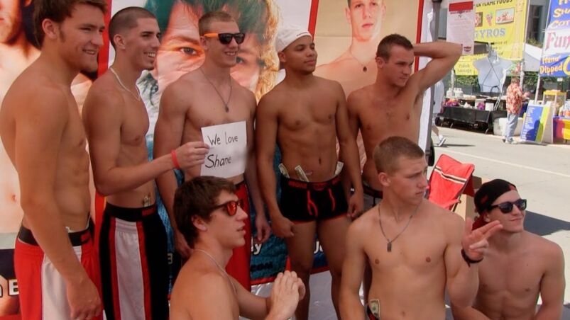 A group of straight men pose shirtless outside in front of a promotional backdrop for their adult film studio.