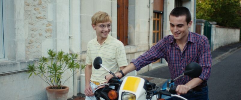 Two young men walk the streets of a small Parisian town while pushing a yellow motorbike between the,