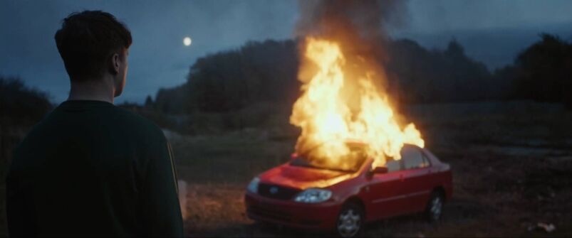Outside at night, we see the back of a man's head as he looks on at a burning car.
