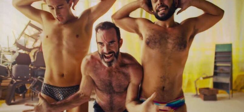 Three shirtless men wear speedos and dance in the middle of a sparse, yellow apartment.