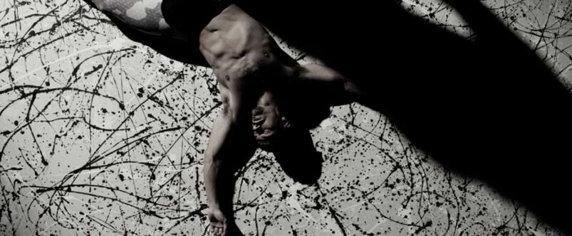 In black-and-white, a shirtless man lays on the splatter-patterned ground, cast in shadow.