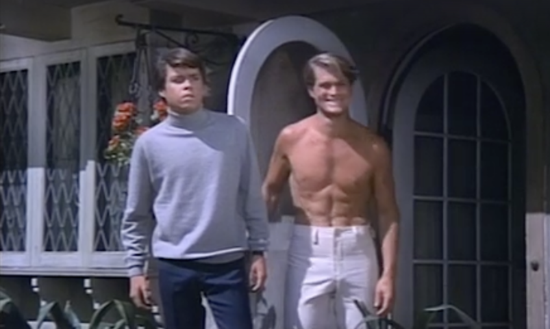 Two men stand outside their front door on a sunny day. The man on the right has brown hair and wears a turtleneck; the man on the left is blond, shirtless, and wearing white pants
