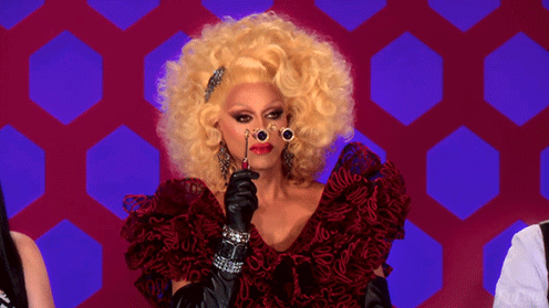 Drag queen RuPaul in a scarlet red dress, black gloves, and a blonde wig, holding tiny opera glasses up to her face.