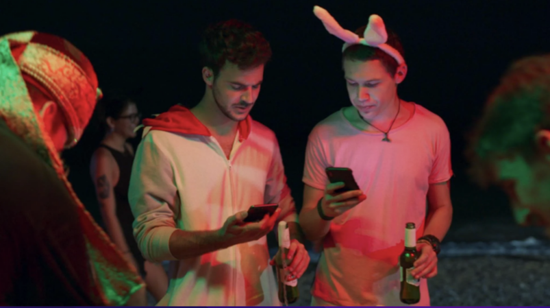 Two men stand beside each other at a dark party looking at their phones. One is wearign a white zip-up, while the other wears costume bunny ears