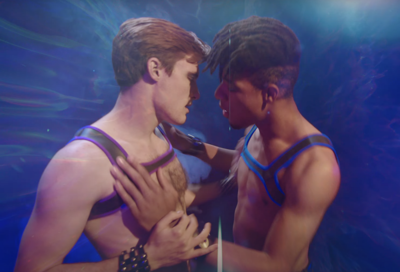 Two fit young men, wearing some sort of fashion harness pieces, lean in for a kiss, surrounded by a light blue fog. 